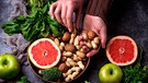 Frau hält Nüsse in den Händen über einer Schale mit Walnüssen und Erdnüssen, daneben eine aufgeschnittene Grapefruit und Brokkoli, Spinat und Äpfel | Bild: mauritius images / Yuliya Furman / Alamy / Alamy Stock Photos
